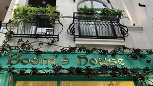 Ein Gebäude mit mehreren Balkonen, die mit Pflanzen und Blumen geschmückt sind. Der Eingang weist ein Schild mit der Aufschrift "Boom's House" in einem türkis gefassten Rahmen auf.