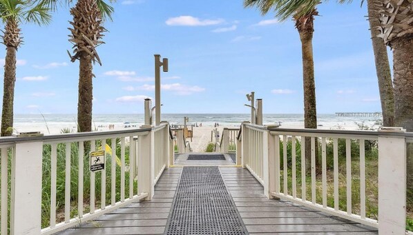 A wooden boardwalk leading to a beach with palm trees and a pier in the background.