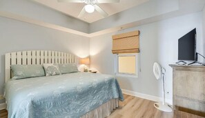 Une chambre à coucher avec un lit, une lampe de chevet, une fenêtre avec un store, un ventilateur de plafond et un téléviseur posé sur un meuble.