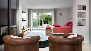 Ein modernes Schlafzimmer mit einem Bett, zwei braunen Ledersesseln, einem kleinen Tisch und Blick auf den Garten durch eine Schiebetür.