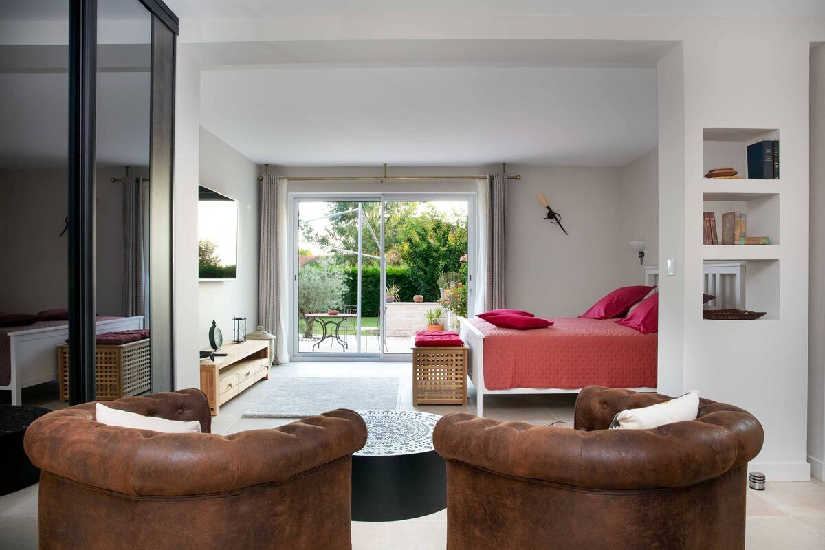 A modern bedroom with a bed, two brown leather armchairs, a small table, and a view of the garden through a sliding glass door.