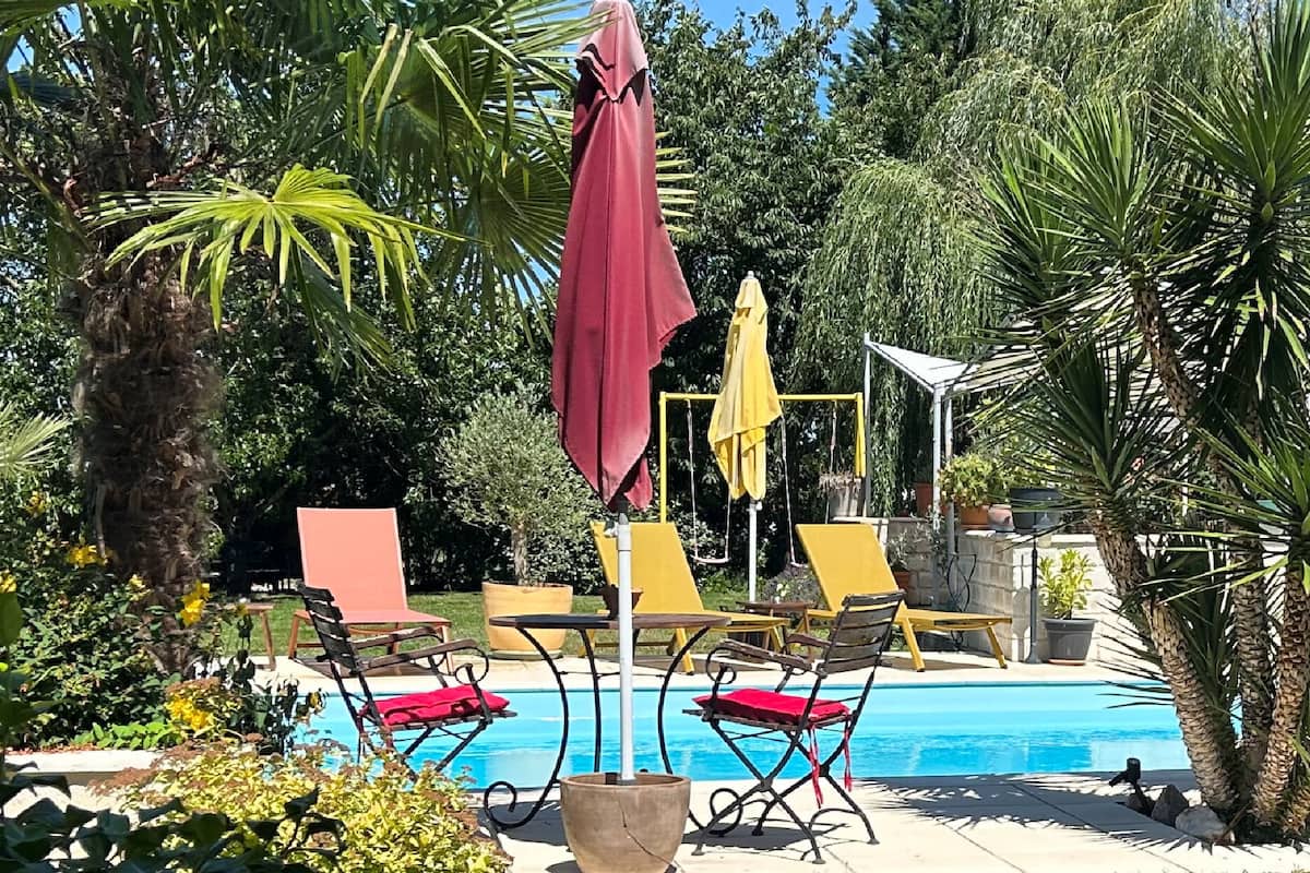 A poolside area with lounge chairs and umbrellas, surrounded by tropical plants and a clear blue pool.