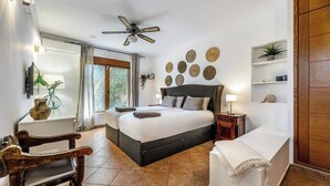 A bedroom with a large bed, a wooden chair, a bedside table, a nightstand, a ceiling fan, a window with curtains, and a wall with decorative plates.