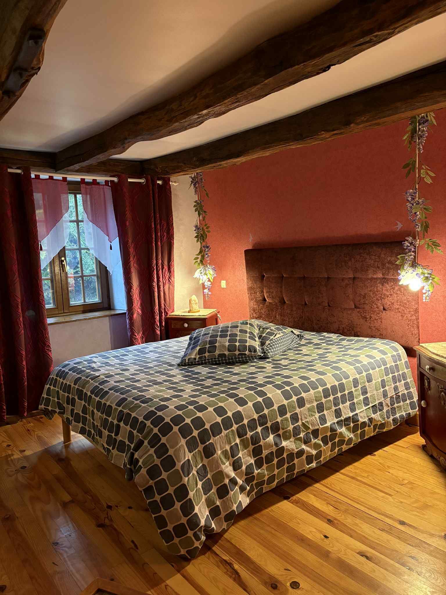 Un dormitorio con piso de madera, una cama con cubrecama estampado, cabecera, mesita de noche, ventana con cortinas y una planta montada en la pared.