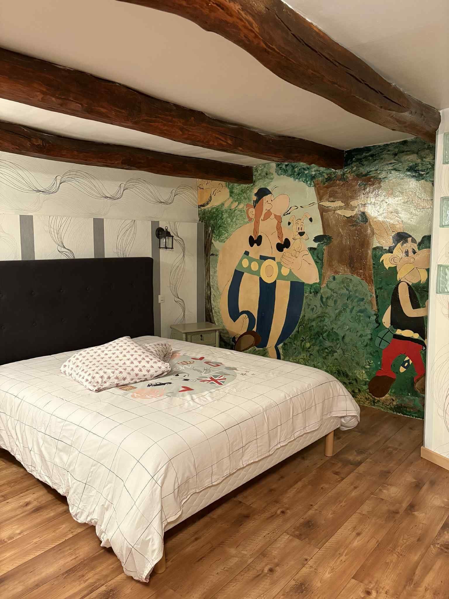 A bedroom with a large bed, wooden beams, a mural, and a nightstand.