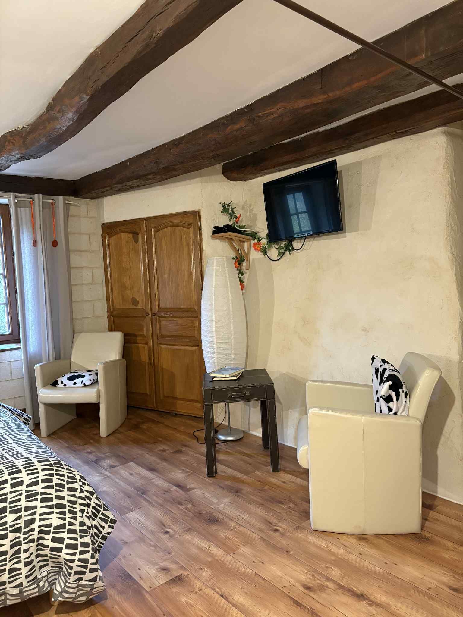 Une pièce avec des poutres apparentes, un téléviseur fixé au mur, une armoire en bois, deux chaises, une petite table et une couvre-lit à motifs.
