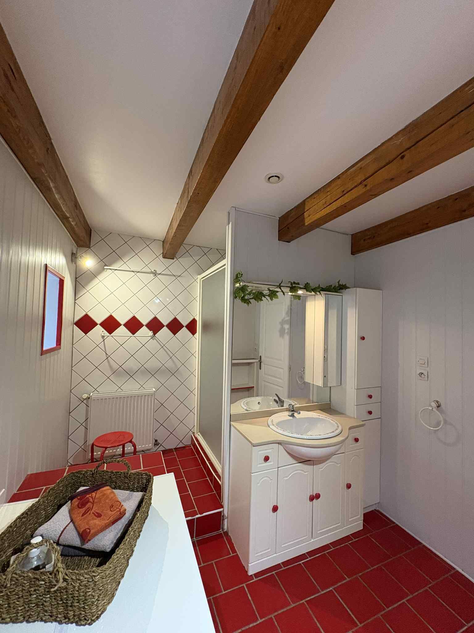 Baño con lavamanos, espejo y muebles blancos, suelo de baldosas rojas y vigas de madera en el techo.