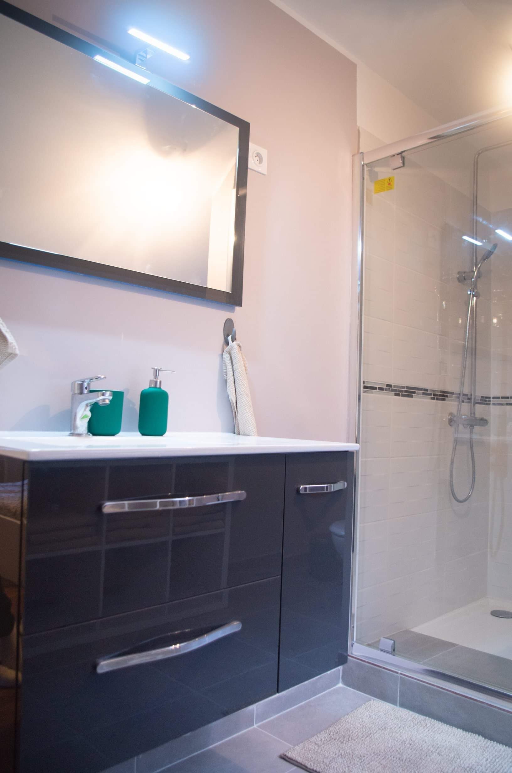 Ein modernes Badezimmer mit Duschkabine aus Glas, einem weißen Waschbecken mit grünem Seifenspender und einem dunklen Waschtisch mit silbernen Griffen.