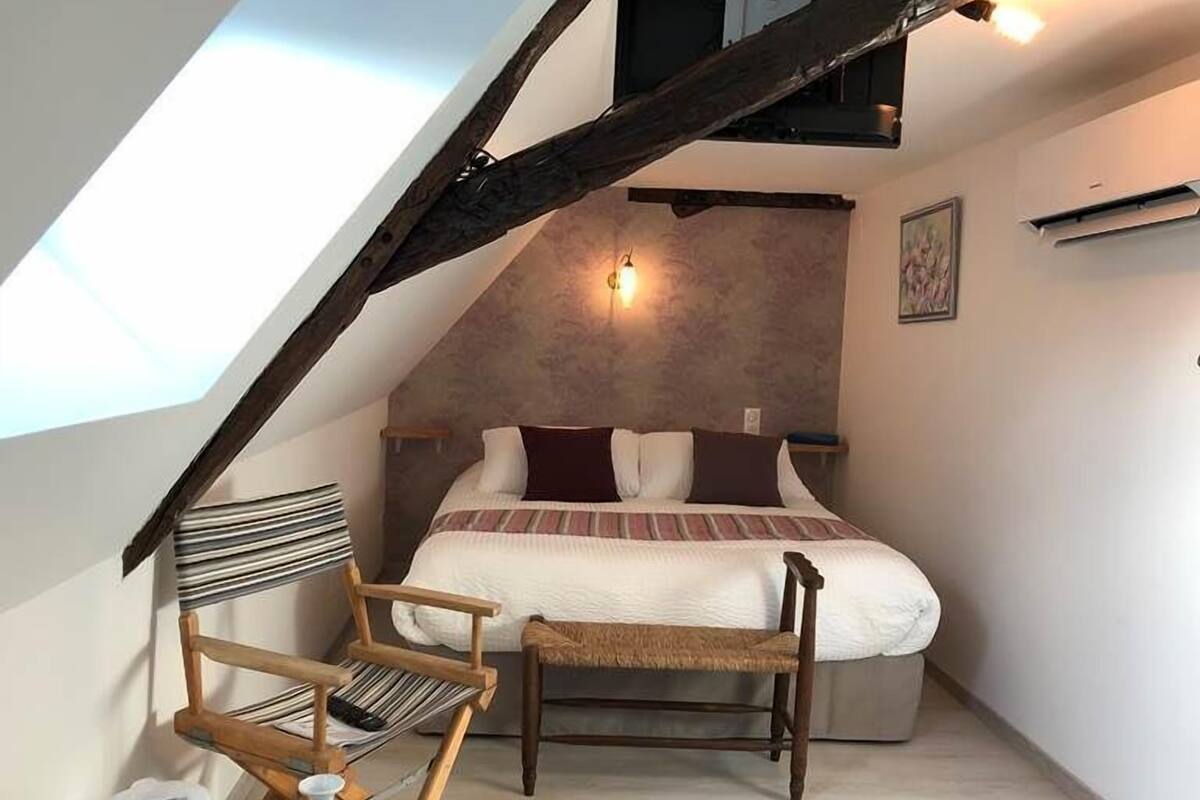 Une chambre avec un plafond en pente, un lit avec une literie à rayures, une chaise en bois et une petite table sur laquelle se trouve une bouteille.