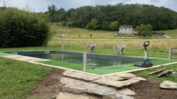 Un chemin de pierres mène à une piscine rectangulaire bordée de verdure, elle-même entourée de pelouse et d’une zone boisée.