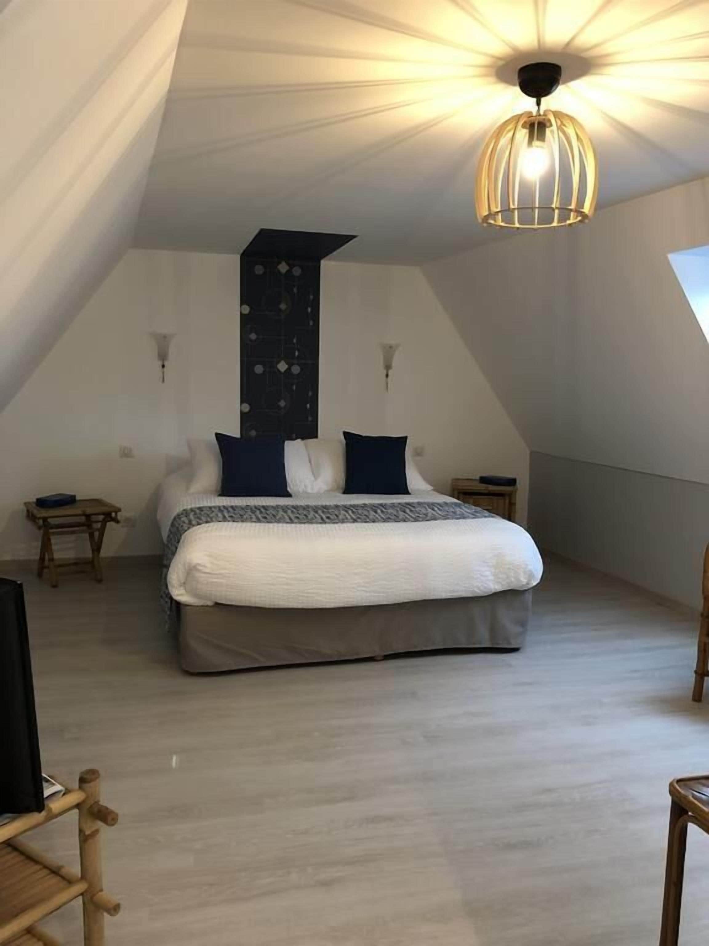 A bedroom with a sloped ceiling, a bed with a white and gray duvet, two pillows, a bedside table, and a hanging light fixture.