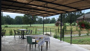 Een terras buiten met een rieten dak, stenen vloer en een tafel met stoelen.