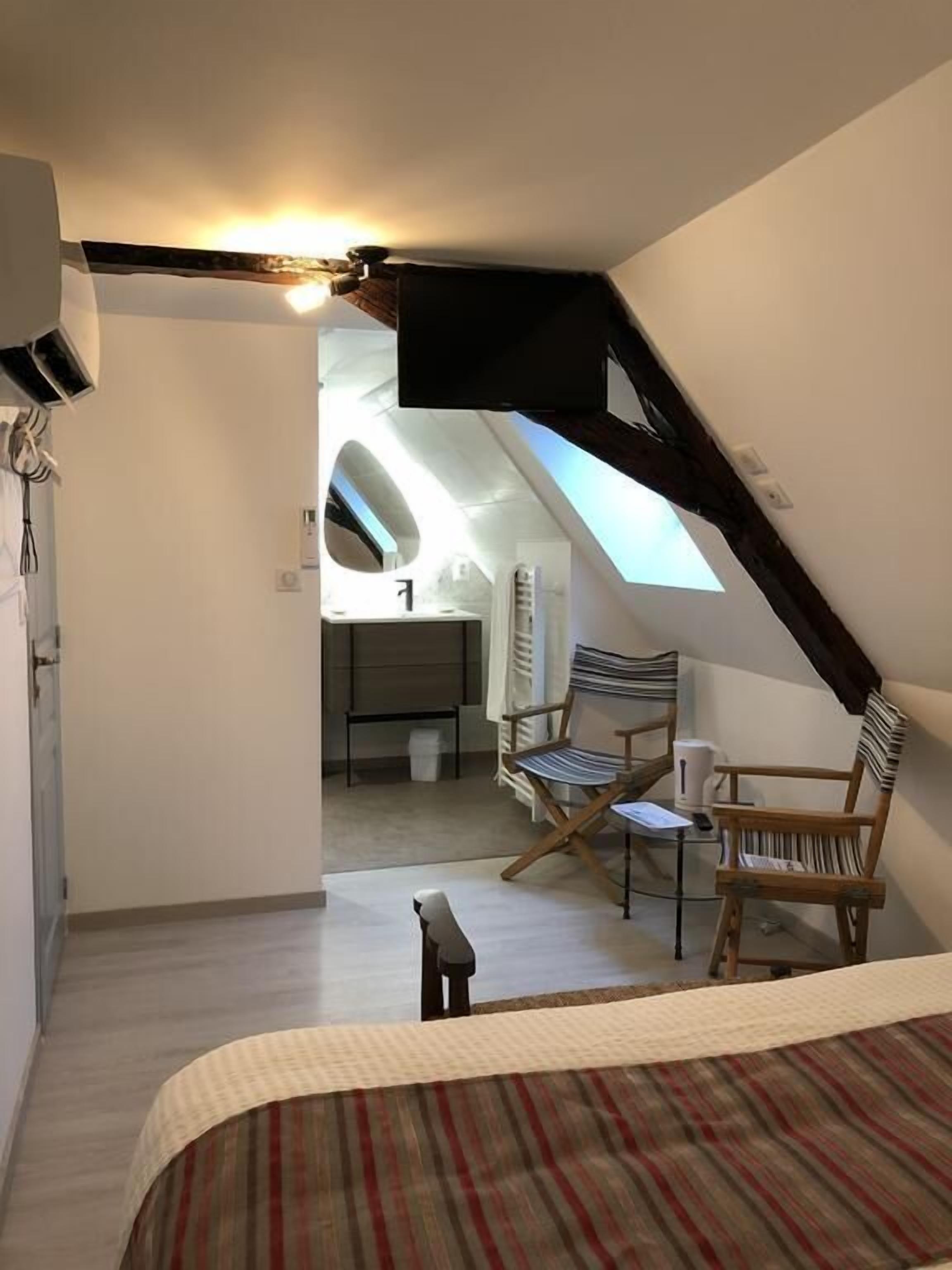 Un dormitorio con techo inclinado, una cama, dos sillas y una pequeña mesa. Se ve un baño al fondo.