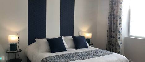 A neatly made bed with a white and blue patterned bedspread, two matching pillows, and a dark headboard. There are two bedside tables with lamps, and a window with patterned curtains.
