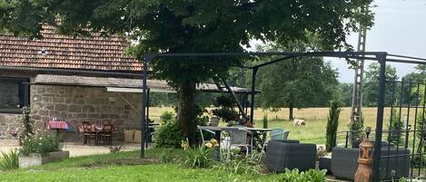 A rustic stone building with a tiled roof, a covered outdoor seating area with chairs and a table, and a large tree providing shade.