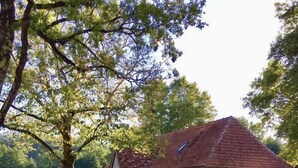 Une maison de campagne en pierre, avec un toit recouvert de tuiles rouges, située au bord d’un plan d’eau paisible, et entourée de verdure et d’arbres.