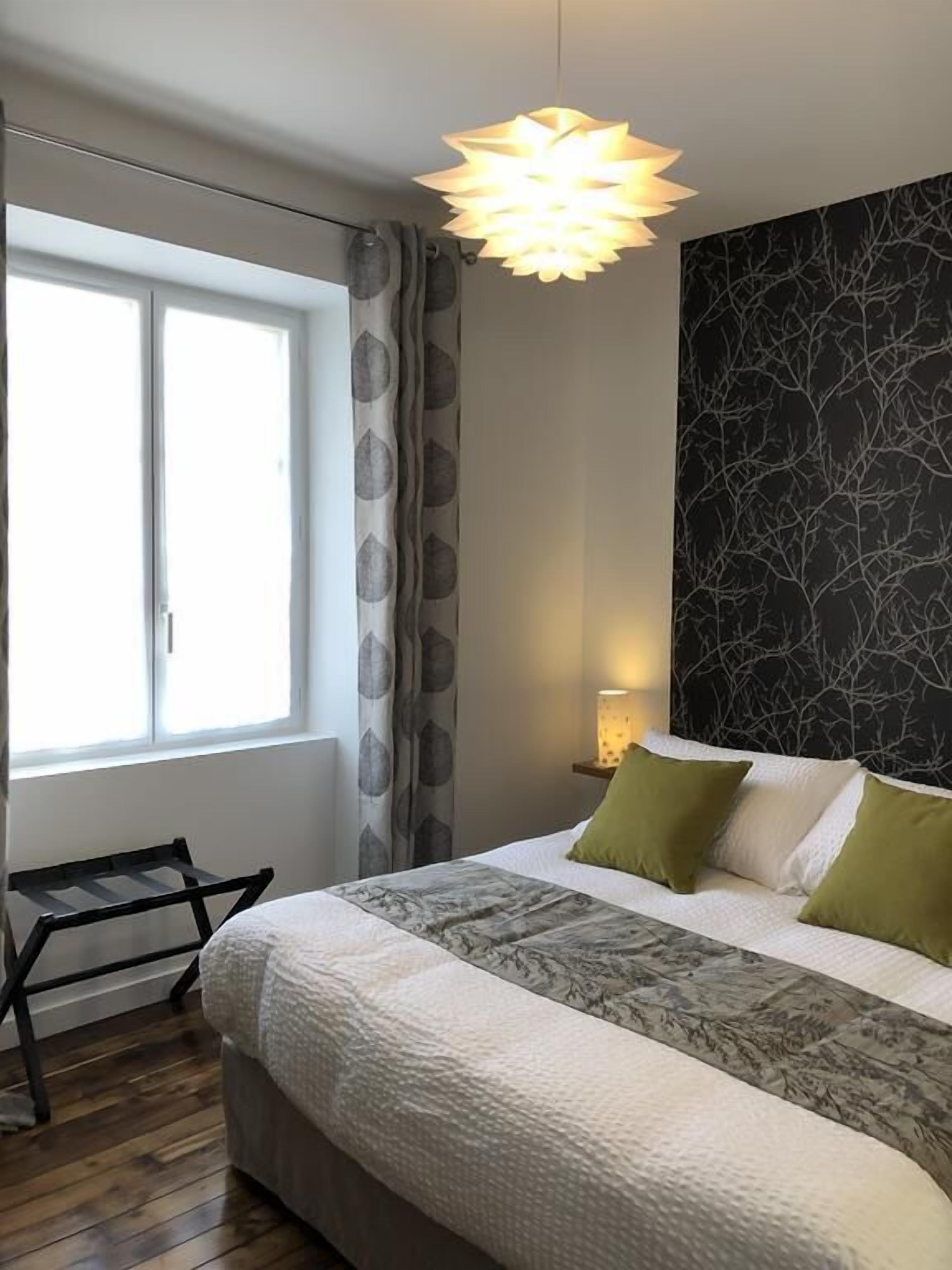 Une chambre à coucher avec un lit, une fenêtre avec des rideaux, une table de chevet avec une lampe et un mur orné d’un motif d’arbre.