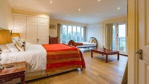 A spacious bedroom with a large bed, a sitting area with a bench, and a view of the outdoors through large windows. - The Homestead by Bloom Stays - sleeps 17 (Canterbury)