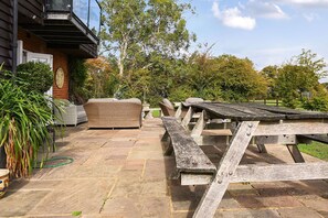 Een terras buiten met stenen vloer, rieten meubels en een houten picknicktafel.