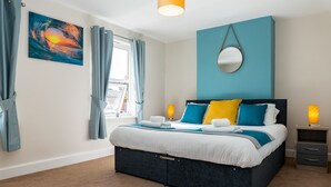 A bedroom with a bed, curtains, a window, a wall-mounted mirror, and a bedside table with a lamp. - Comfy, 2 Bed House, Sleeps 6 with Garden and Free Parking in Cheltenham (Cheltenham)
