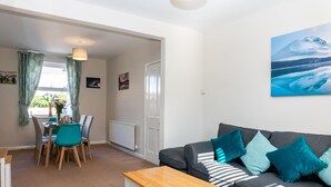 A modern living room with a grey sofa, teal cushions, a wooden coffee table, and a dining area with a table and chairs. - Comfy, 2 Bed House, Sleeps 6 with Garden and Free Parking in Cheltenham (Cheltenham)