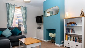 A living room with a grey sofa, a wooden coffee table, a white bookshelf, a flat-screen TV, and a blue accent wall. - Comfy, 2 Bed House, Sleeps 6 with Garden and Free Parking in Cheltenham (Cheltenham)