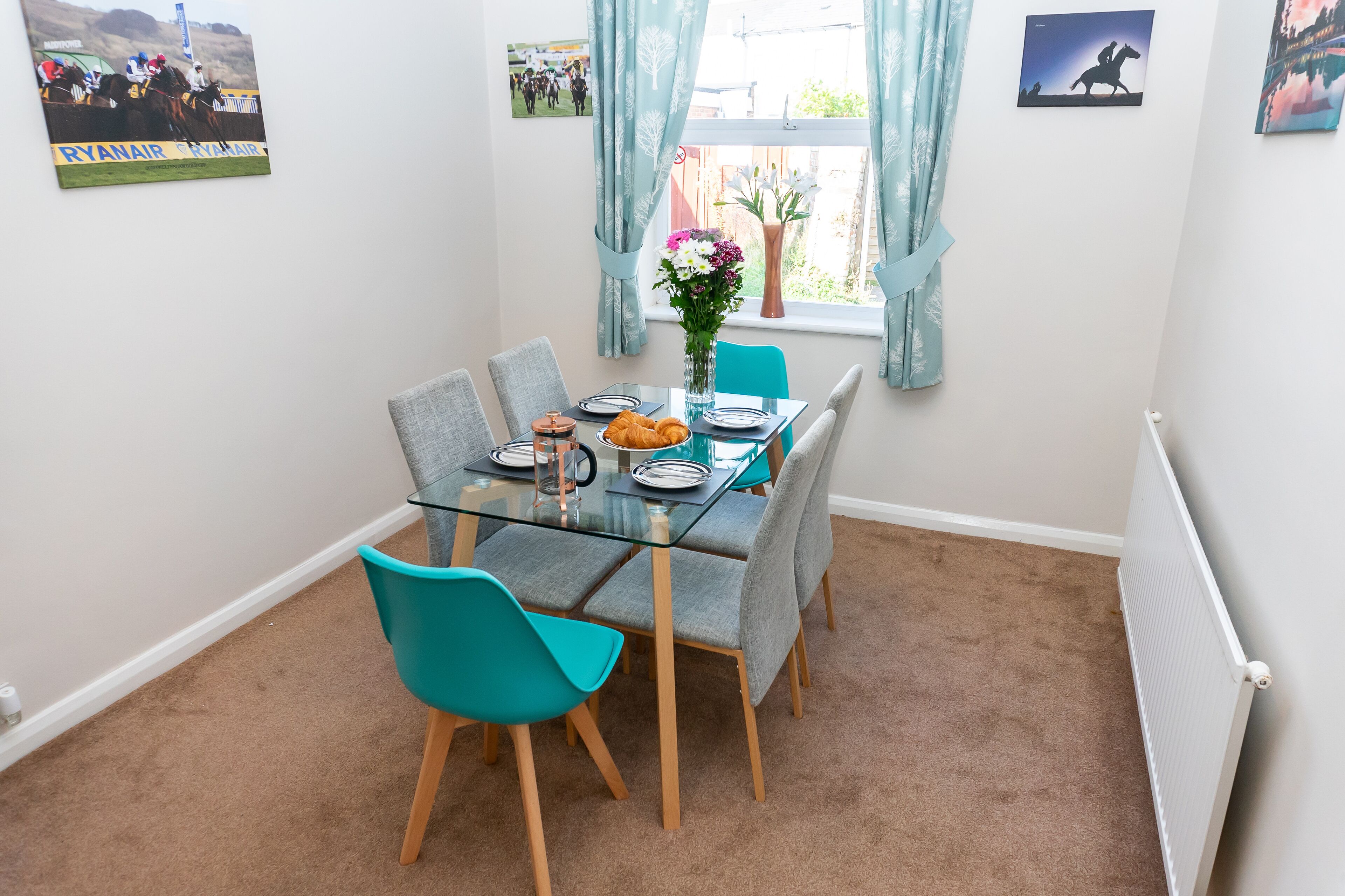 Een eethoek met een glazen tafel, teal stoelen en een bloemenvaas.