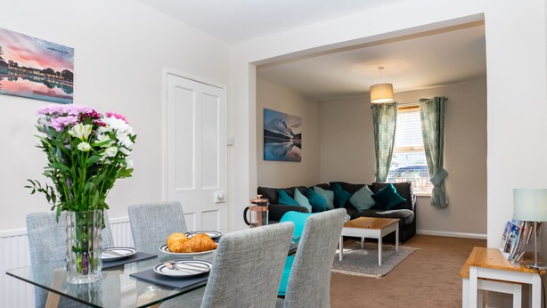 A dining area with a glass table, grey chairs, a vase of flowers, and a croissant on the table. - Comfy, 2 Bed House, Sleeps 6 with Garden and Free Parking in Cheltenham (Cheltenham)