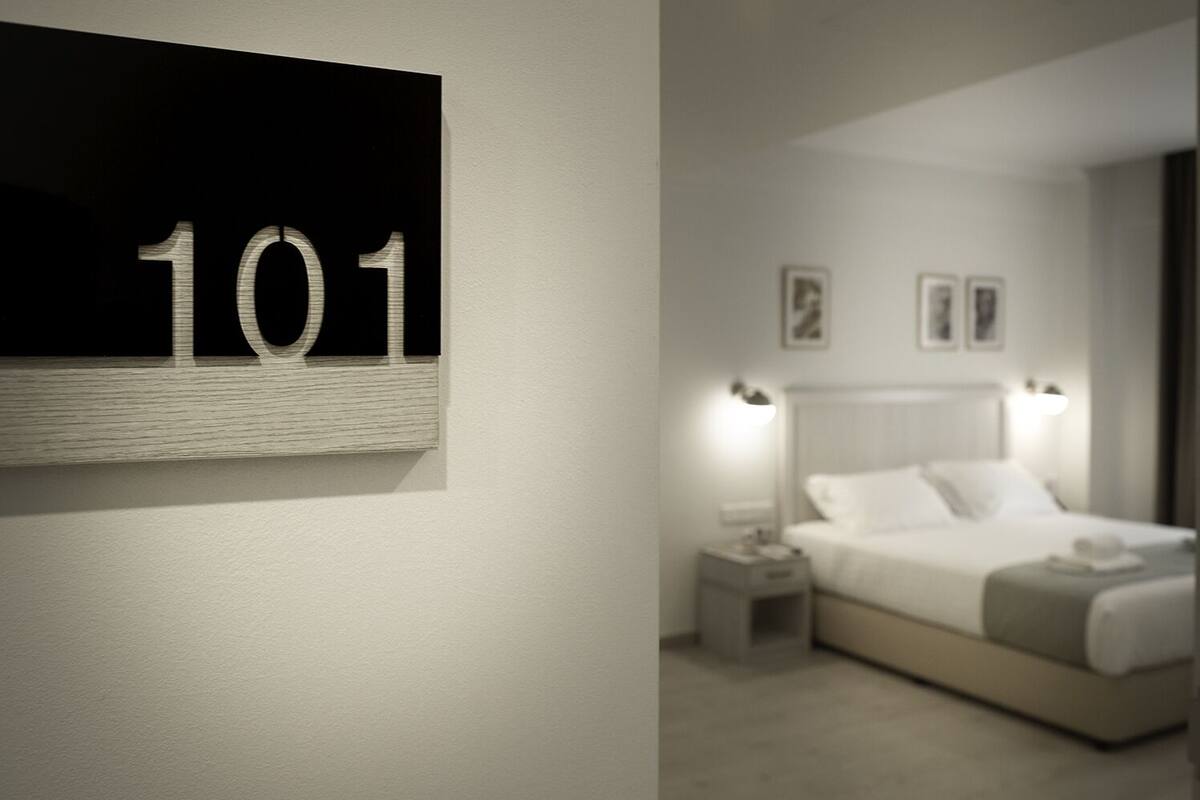 A hotel room with a large number '101' on the wall, a neatly made bed with white linens, two bedside lamps, and two framed pictures on the wall.