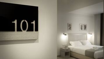 A hotel room with a large number '101' on the wall, a neatly made bed with white linens, two bedside lamps, and two framed pictures on the wall.