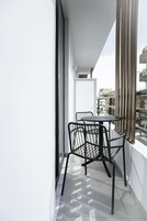 Uma varanda com mesa redonda e quatro cadeiras, uma porta branca e vista para edifícios através de janelas amplas.