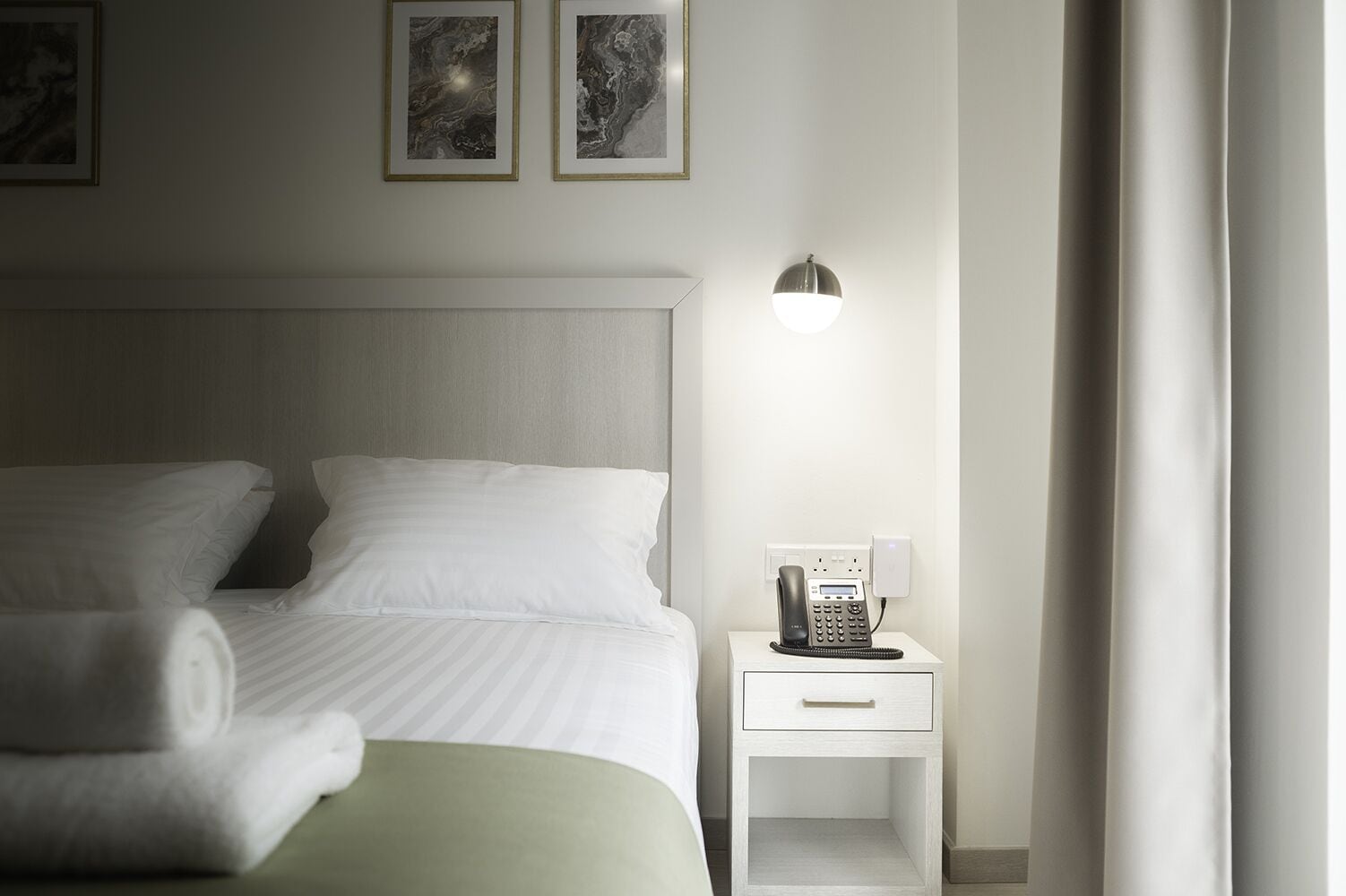Cama arrumada com lençóis brancos, mesa de cabeceira com telefone e luminária fixada na parede.