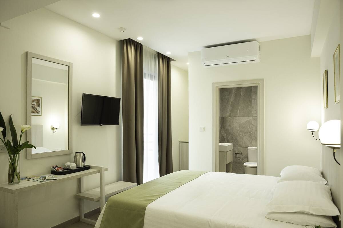 A hotel room with a bed, a desk with a flat-screen TV, a mirror, a vase with flowers, and an air conditioning unit.
