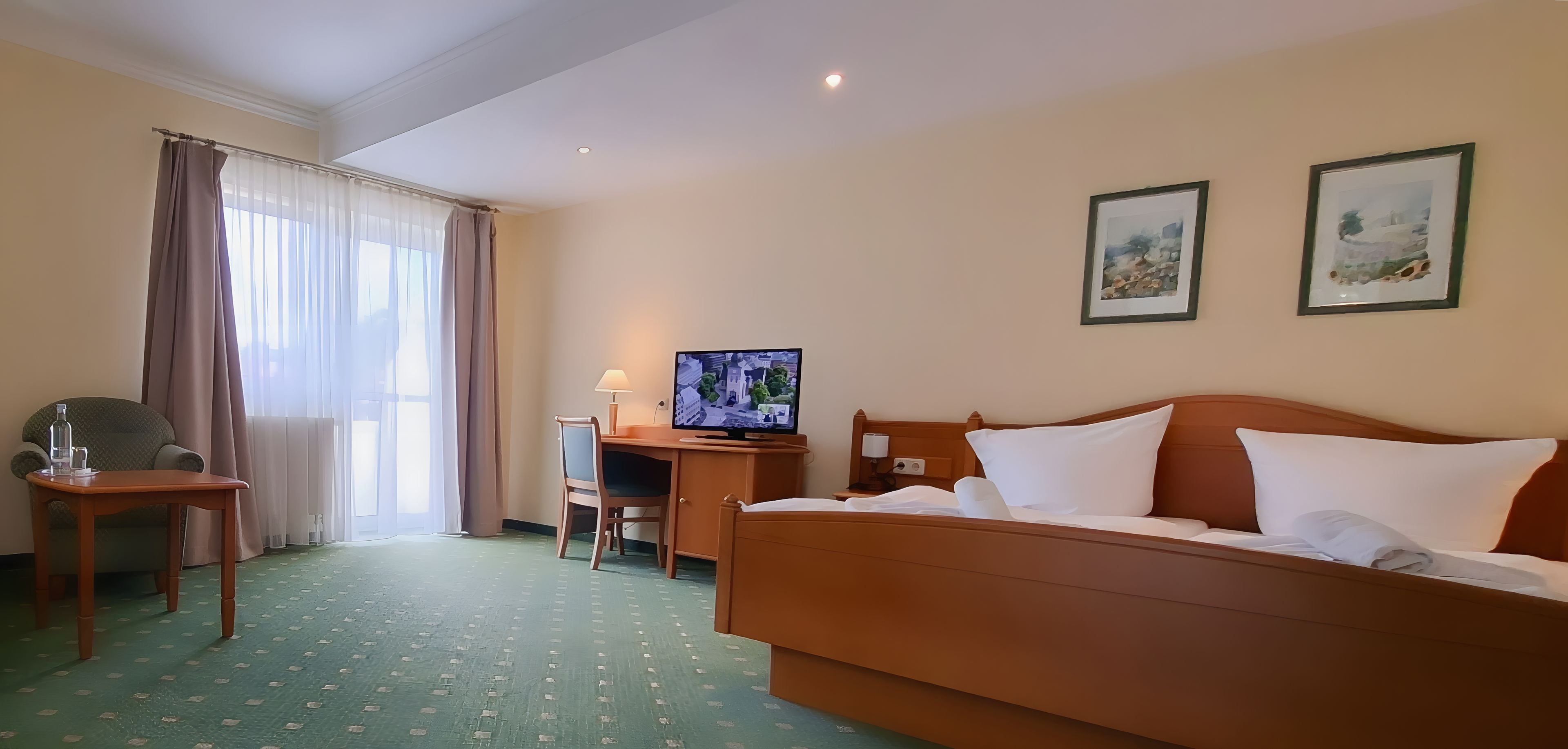 A hotel room with a bed, a desk with a computer, a chair, a window with curtains, and two wall-mounted pictures.