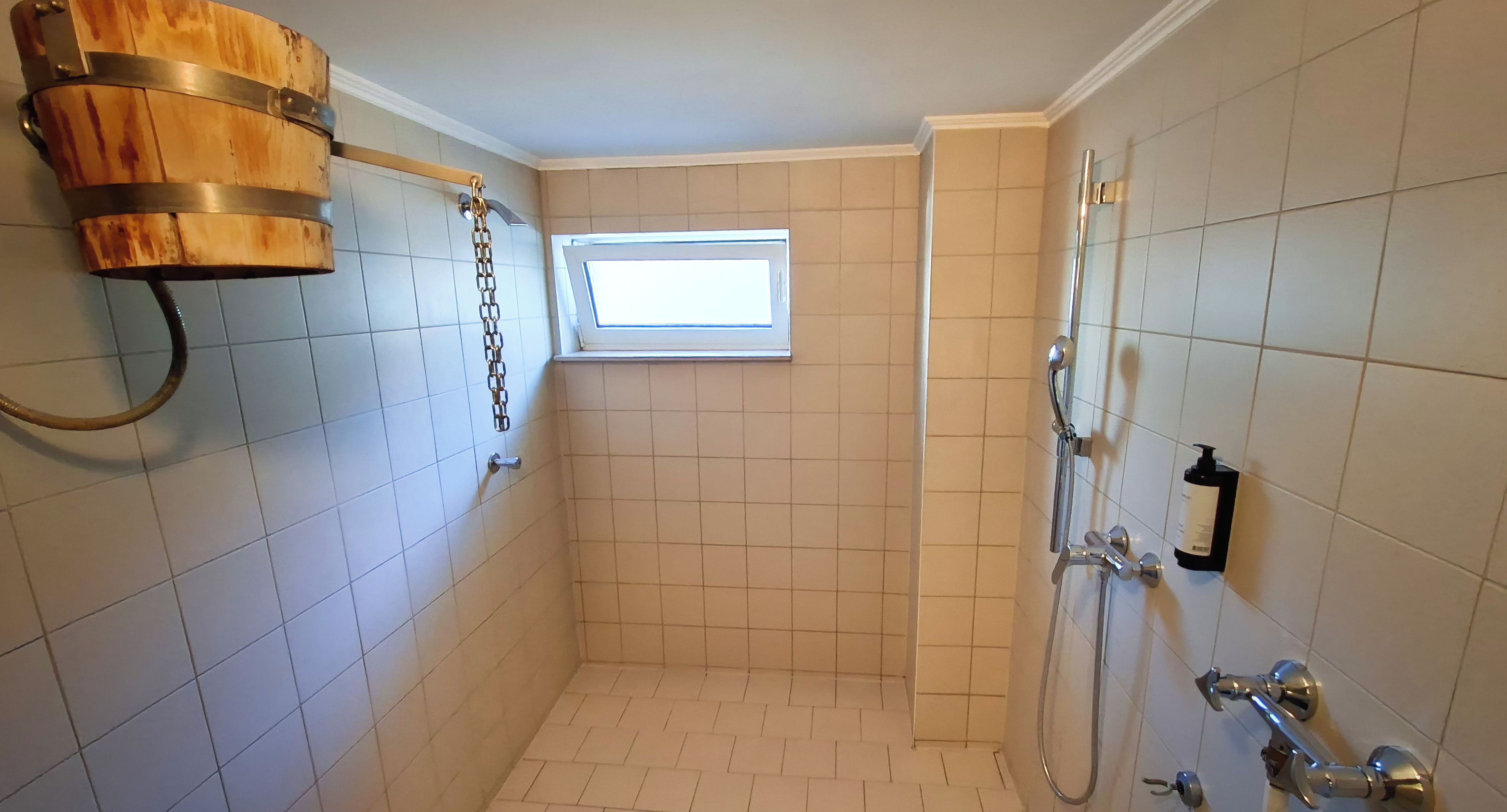 A small, tiled shower room with a wooden bucket, a window, and a showerhead.