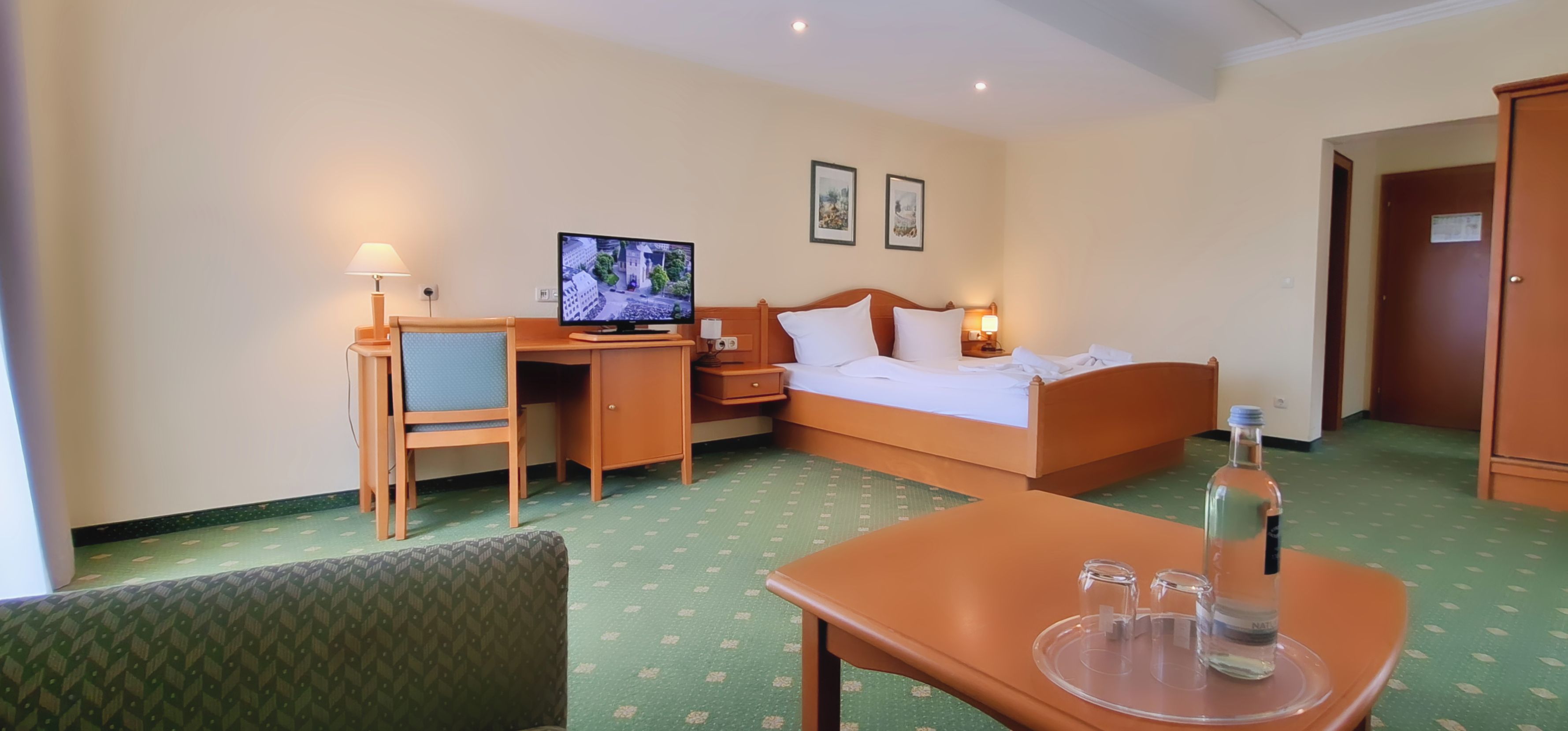 Una camera d'hotel con un letto, una scrivania, una sedia, televisione e un piccolo tavolo con bicchieri e una bottiglia.