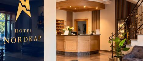 A hotel reception area with a wooden reception desk, a large mirror, and a staircase with ornate railings.