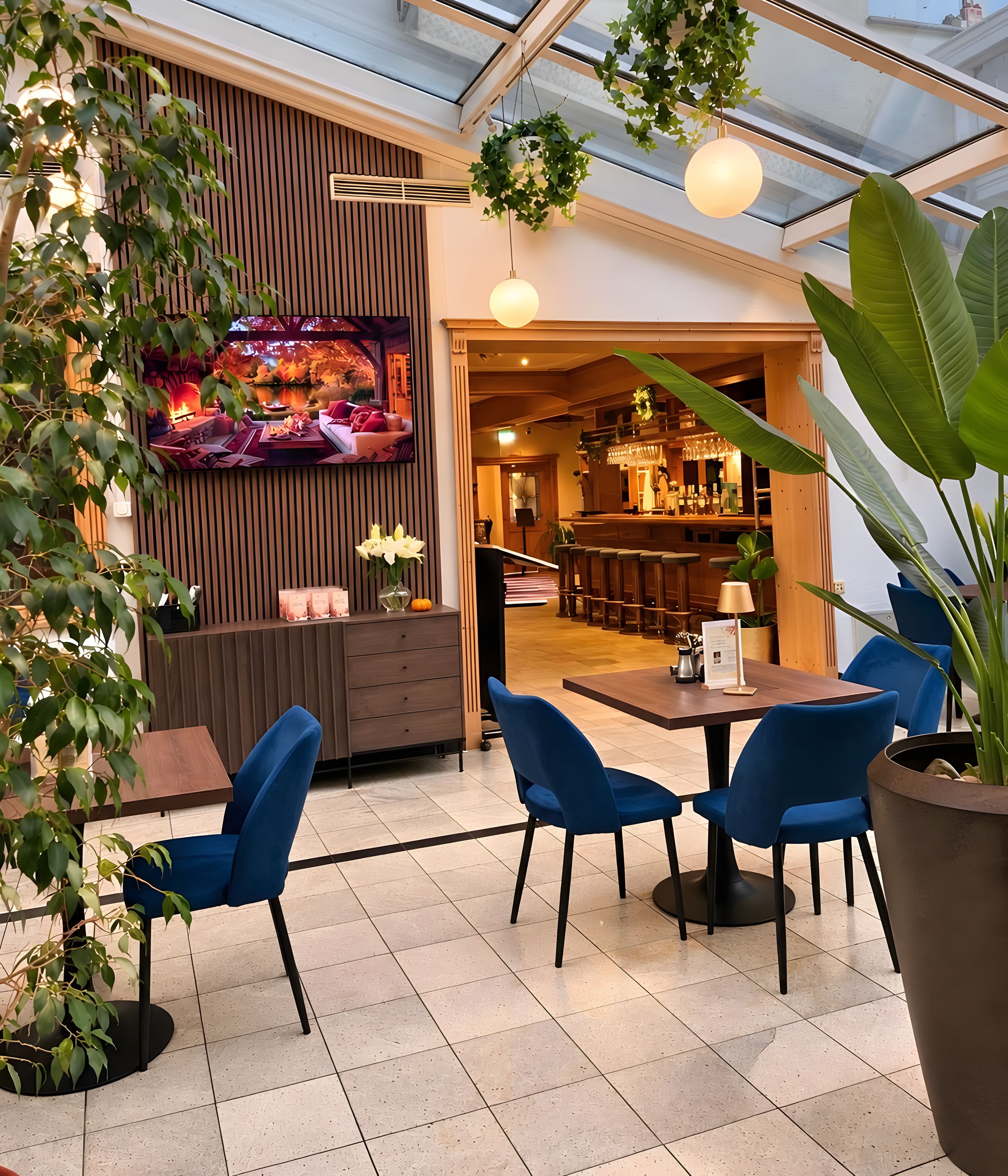 Un espace intérieur moderne avec un coin repas comprenant des chaises bleues et une table en bois, le tout agrémenté de plantes en pot et d’un grand téléviseur à écran plat.