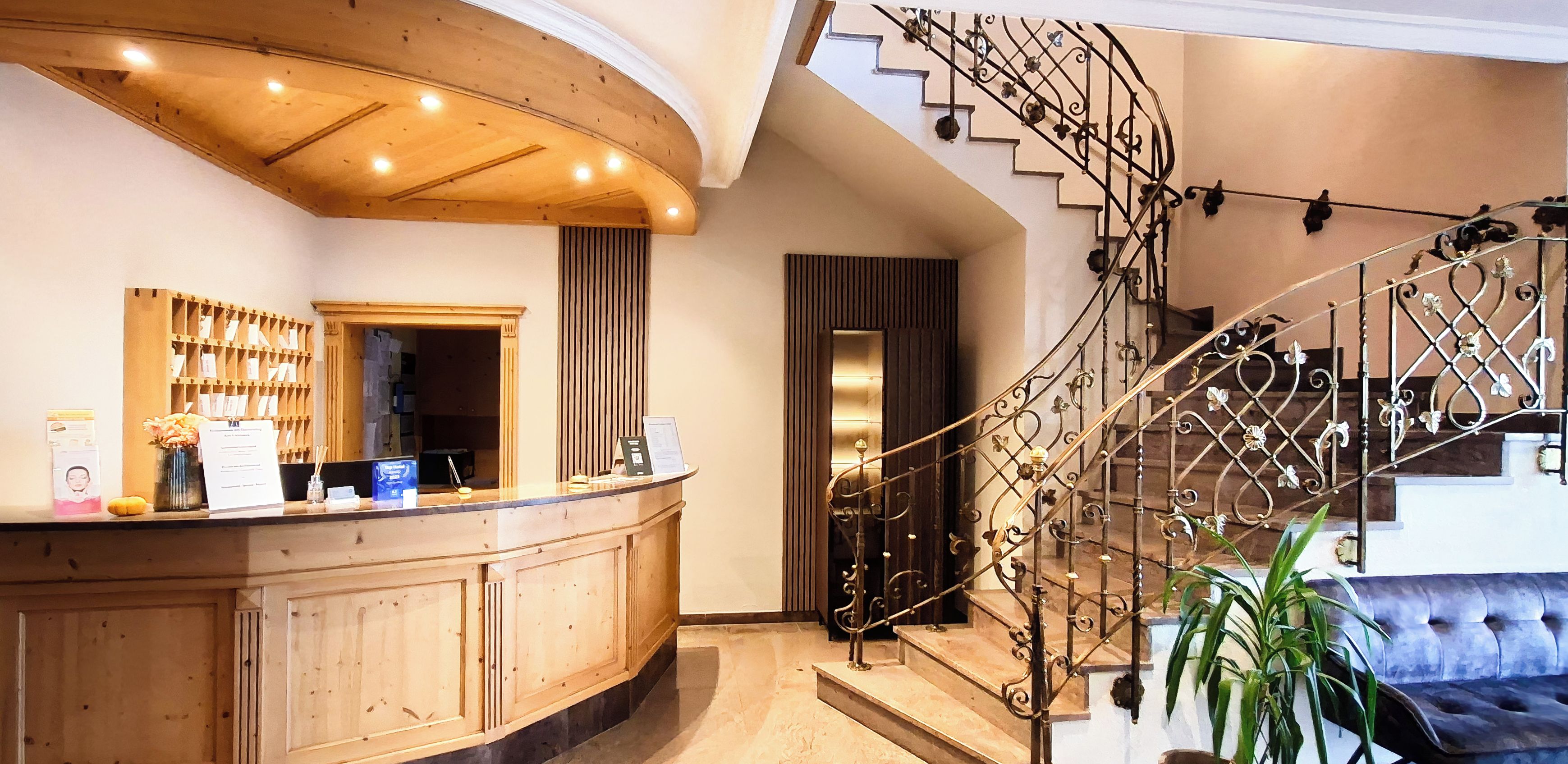 Una recepción con mostrador de madera, una escalera con barandilla de metal ornamentada y un sofá tapizado en azul.