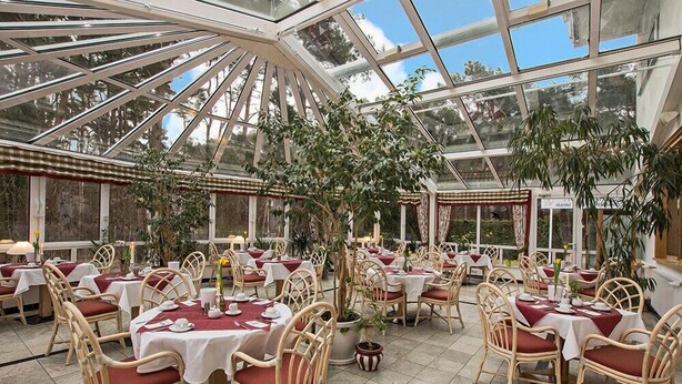 A conservatory with tables and chairs, large windows, and potted plants. - Hotel Nordkap (Karlshagen)