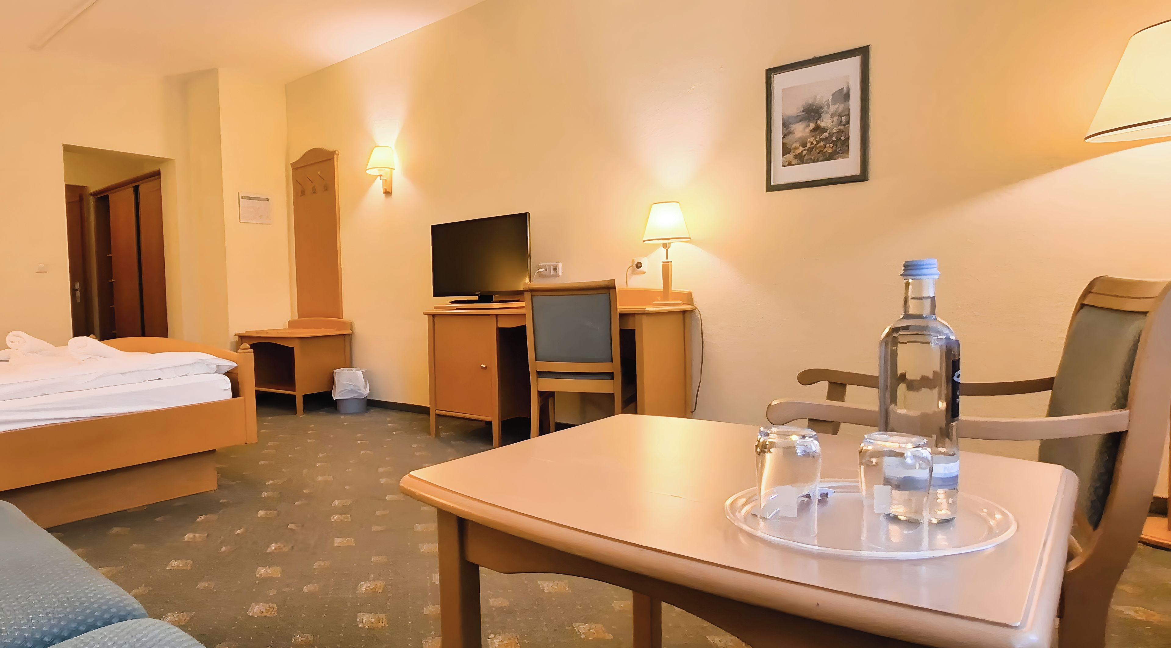 A hotel room with a bed, a desk, a chair, a TV, a lamp, and a table with a bottle and glasses.