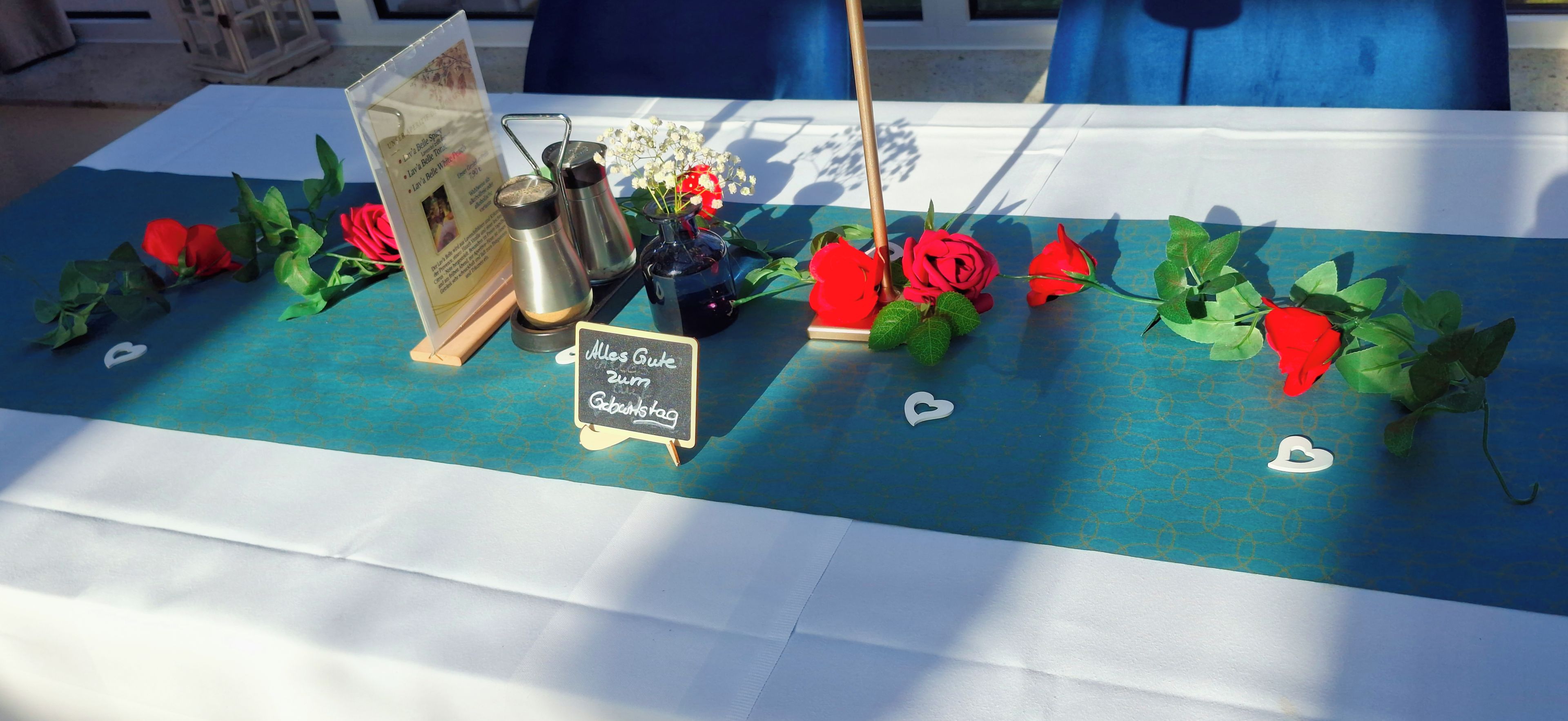 A table with a blue tablecloth, red roses, a small sign, and a coffee maker.