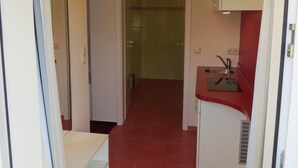 Een smalle gang met een vloer van rode tegels, een kleine kitchenette met een gootsteen en fornuis, en een glazen douchedeur.
