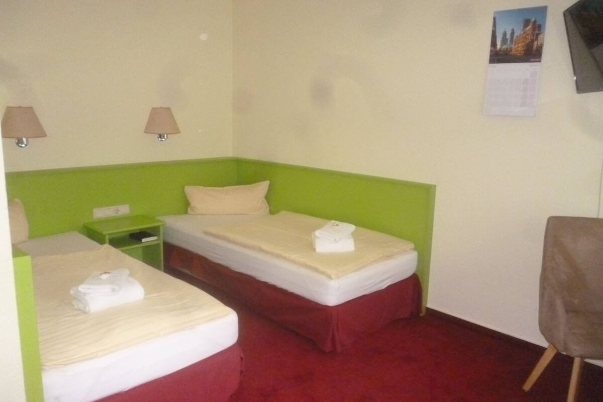 A hotel room with two single beds, a green headboard, a red carpet, and a small table with a lamp.