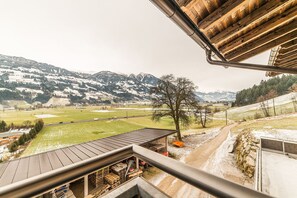 Vista desde un balcón sobre un paisaje rural con montañas nevadas, un sendero de tierra y una construcción de madera con techo de metal.