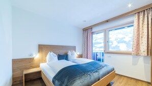 A bedroom with a wooden bed frame, white bedding, and a view of a snowy landscape through a window. - Aberghof Apartments (Fuegen)
