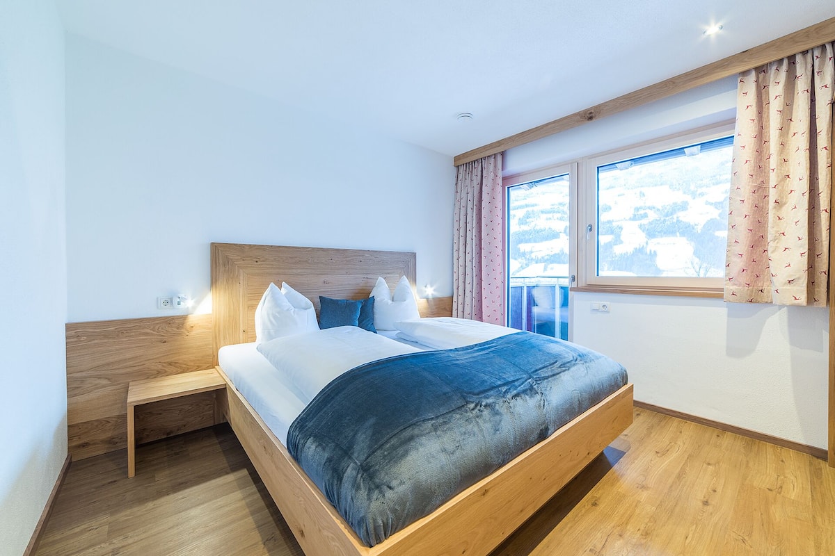 Un dormitorio con estructura de cama de madera, ropa de cama blanca y vista a un paisaje nevado a través de una ventana.