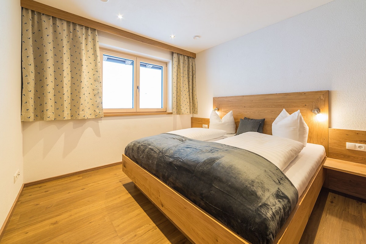 Un dormitorio con estructura de cama de madera, un ventanal con cortinas y una mesita de noche con lámpara.