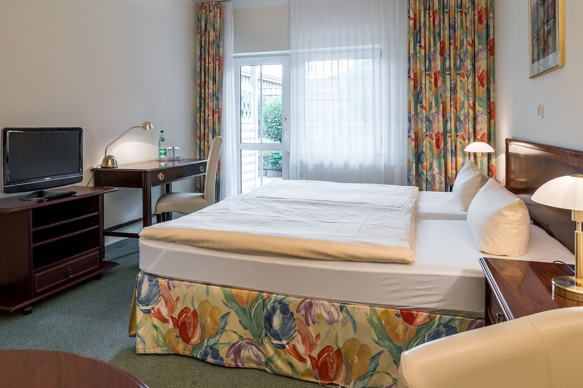 Quarto de hotel com uma cama, uma escrivaninha com computador, uma cadeira, televisão e janela com cortinas.