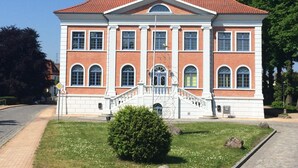 A large, two-story building with a symmetrical facade, featuring a central entrance with a staircase, large windows, and a red-tiled roof.